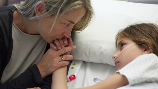 A woman holds a young girl's hand as she lies in a hospital bed with an oxygen tube.